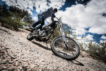 Person standing on a motorcycle in a rocky desert landscape under a cloudy sky.