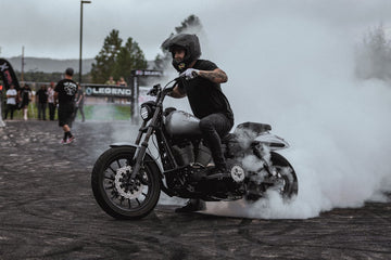 A person on a motorcycle performs a burnout, creating clouds of smoke, with an overcast sky and onlookers in the background.