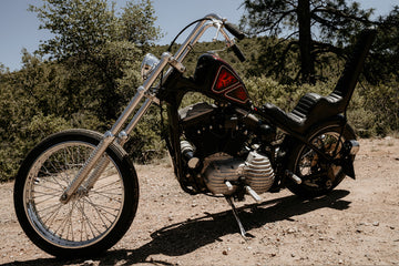 A black custom chopper motorcycle with extended front forks and tall backrest, parked on a dirt path, surrounded by trees and greenery.