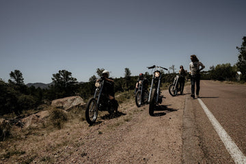 Four people with three motorcycles parked on the side of a rural road, surrounded by trees and a clear sky.