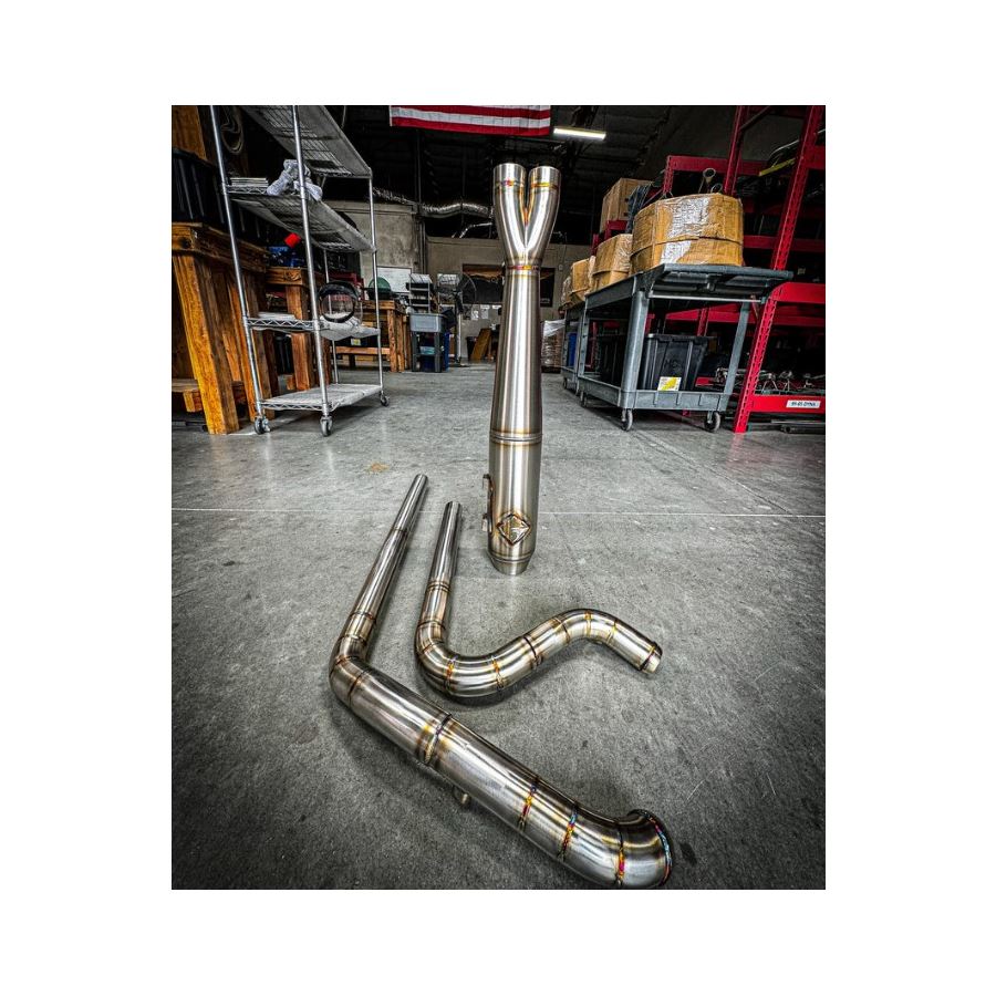 Metal pipes and a Y-shaped exhaust component rest on the concrete floor in a workshop environment, surrounded by shelves and boxesâa scene where SP Concepts works to perfect their Full Length Turnpike Works Edition for Harley &