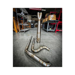 Metal pipes and a Y-shaped exhaust component rest on the concrete floor in a workshop environment, surrounded by shelves and boxesâa scene where SP Concepts works to perfect their Full Length Turnpike Works Edition for Harley '96-'16 Touring exhaust systems.