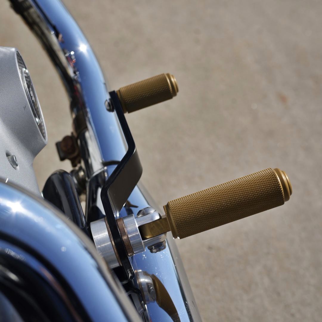A close up of the handlebars on a motorcycle, highlighting the TC Bros. Nomad Foot pegs for Harley Models - Knurled - Gold for added traction and stability.