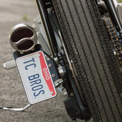 A motorcycle with a license plate attached to it and a TC Bros. Ford Duolamp Model A Stainless Steel Tail Light for the brake/running light.
