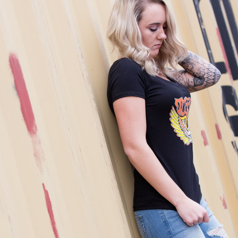A woman wearing a black t-shirt, specifically the TC Bros. Women's Wing Tee - Black from TC Bros., leaning against a wall.