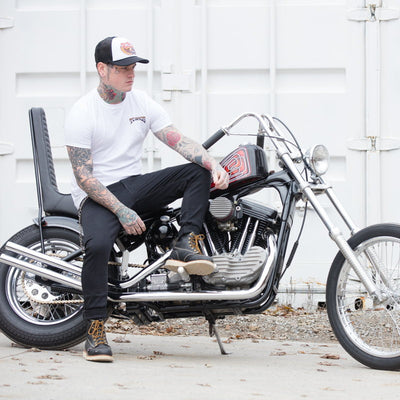 A man with tattoos sitting on a motorcycle wearing a TC Bros. Wing Trucker Hat in White/Black with an adjustable snapback closure.