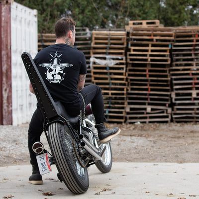 A man, wearing a TC Bros. Ignition T-Shirt - Black, sits on a motorcycle in front of pallets.