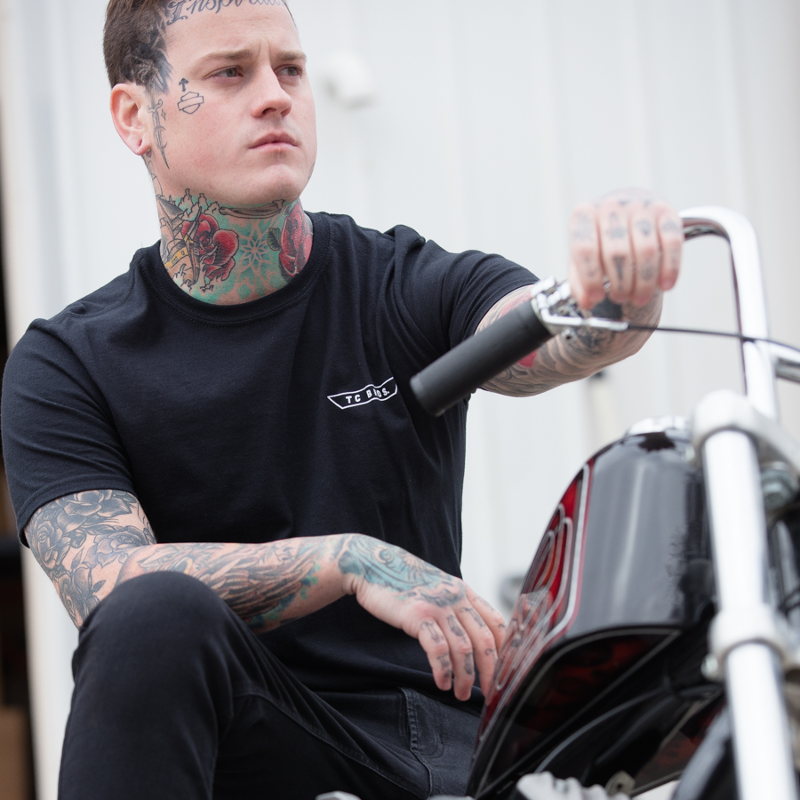 A man with tattoos sitting on a TC Bros. Ignition T-Shirt - Black motorcycle.