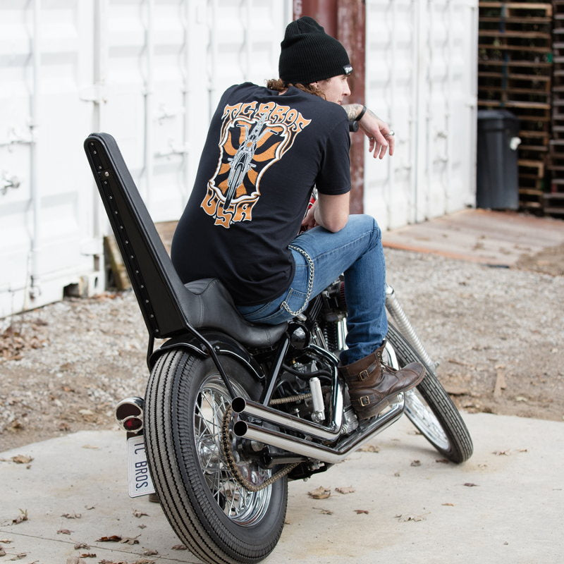 A man wearing a black TC Bros. Trippin' T-Shirt sitting on the back of a motorcycle.