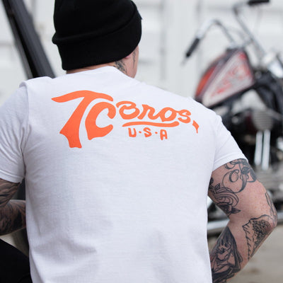 A man wearing a TC Bros. Classic T-Shirt in white with orange lettering.