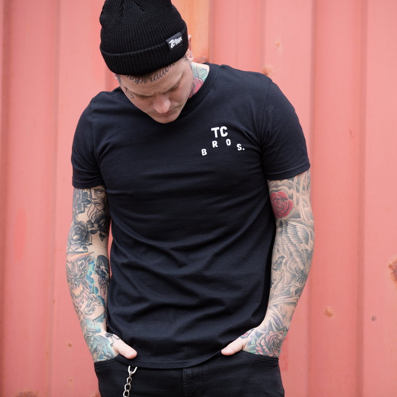 A man wearing a black TC Bros. Devil T-Shirt, adorned with tattoos.