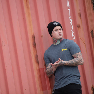 A man with tattoos and a beanie leaning against a wall wearing a TC Bros. Shield T-Shirt in Charcoal Heather.