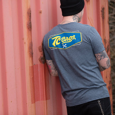 A man wearing a TC Bros. Shield T-Shirt - Charcoal Heather with a yellow and blue logo.