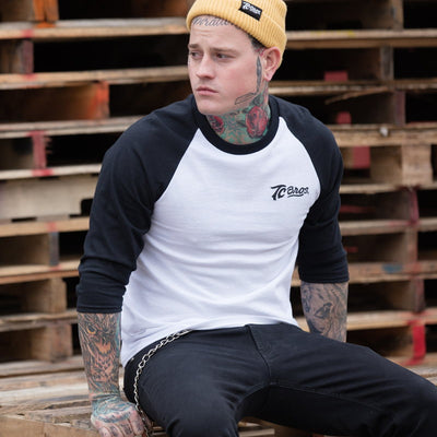 A man with tattoos sitting on a soft acrylic pallet, wearing a TC Bros. Watchman Beanie - Nugget.