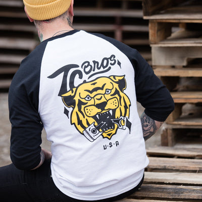 A man sitting on a pallet wearing a TC Bros. Tiger Raglan - White/Black shirt with a tiger on his back.