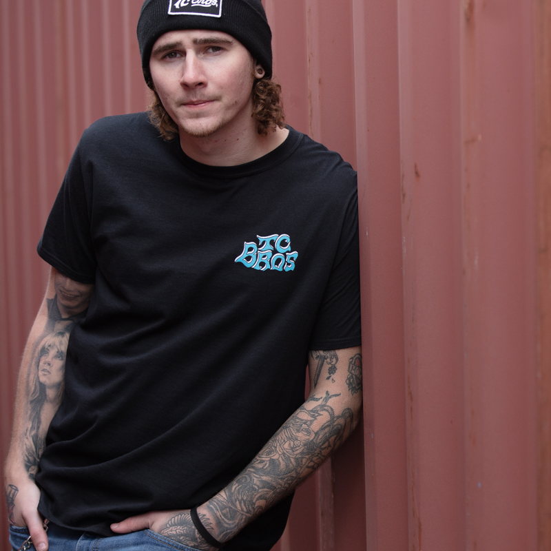 A man wearing a TC Bros. Eagle T-Shirt in Black leaning against a wall.
