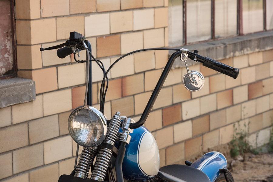 A blue motorcycle parked in front of a brick building, featuring TC Bros. 7/8" Ape Hanger Handlebars - 12" Black Powdercoated made of American steel tubing.