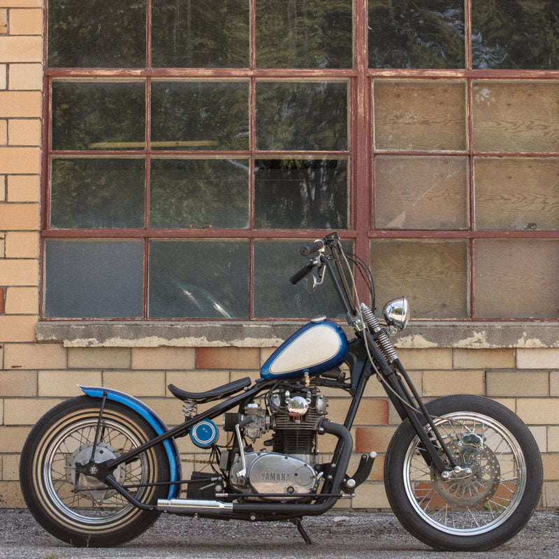 A blue and white TC Bros. Yamaha XS650 Forward Controls Kit motorcycle parked in front of a building.