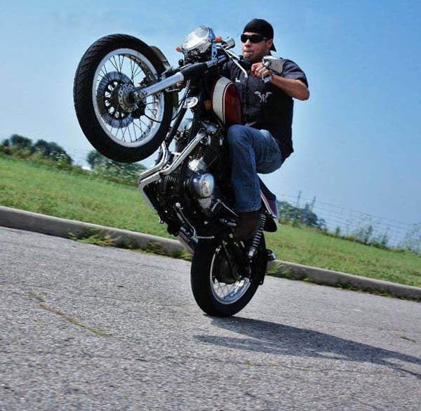A man performing a trick on a [Loaded Gun Customs] motorcycle would be: A man performing a trick on a [Loaded Gun Custom Yamaha XS650 Rearsets by TC Bros.] motorcycle.