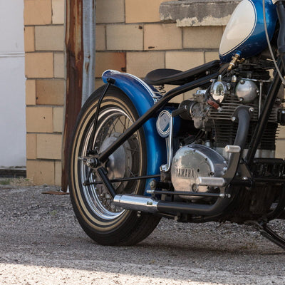 A blue Moto Iron® motorcycle with the 6" Wide Raw Steel Ribbed Bobber Fender parked in front of a brick building.