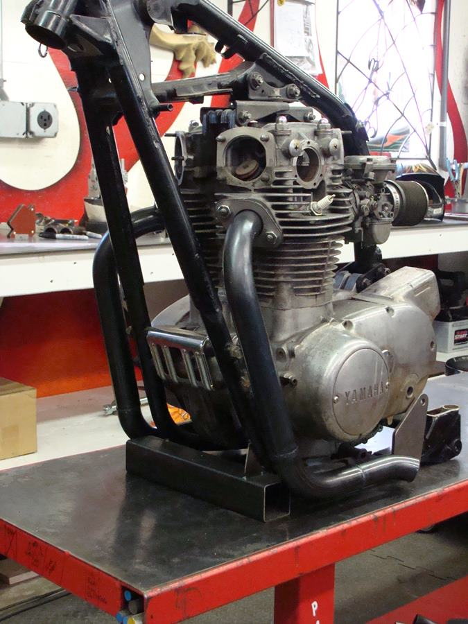A Yamaha XS650 motorcycle engine sitting on a table in a workshop, customized with a Pandemonium "Hot Rod" Exhaust System.