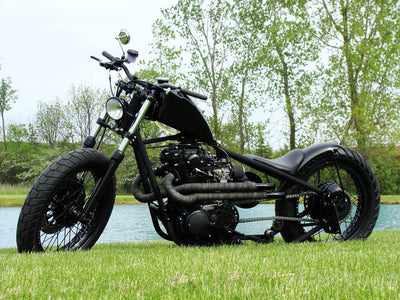 A motorcycle parked in the grass near a lake with a black custom Yamaha XS650 "Ya Mama" Exhaust System By Pandemonium (Fits 1974-1982 XS650).