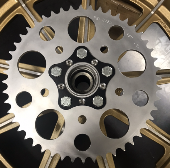 A close-up of a metal sprocket on a gold alloy wheel with bolts and circular cutouts, showcasing the Hugh’s Handbuilt HHB Harley Davidson Sprocket Lock System in black for enhanced security.