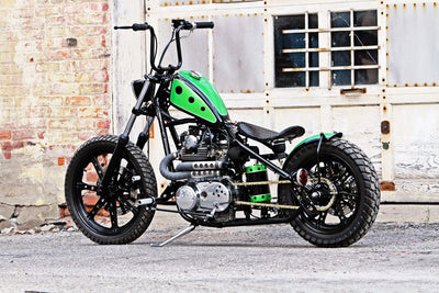A green and black TC Bros. motorcycle with a TC Bros. Chrome Weld On Kick Stand for 1-1/8" Frame Tubing parked in front of a building.