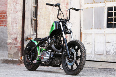 A green and black motorcycle, with a Moto Iron® Triangle Chopper Headlight - Aris Style - Chrome, parked in front of a building.