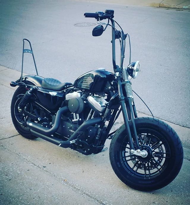 A black motorcycle parked on the side of the road, featuring a Moto Iron® Harley Wishbone Springer Front End for Harley Davidson Dyna 91-17 & Sportster 04-Up (-3" Under, Black).