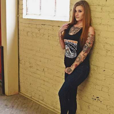 A woman with tattoos leaning against a TC Bros. black brick wall.