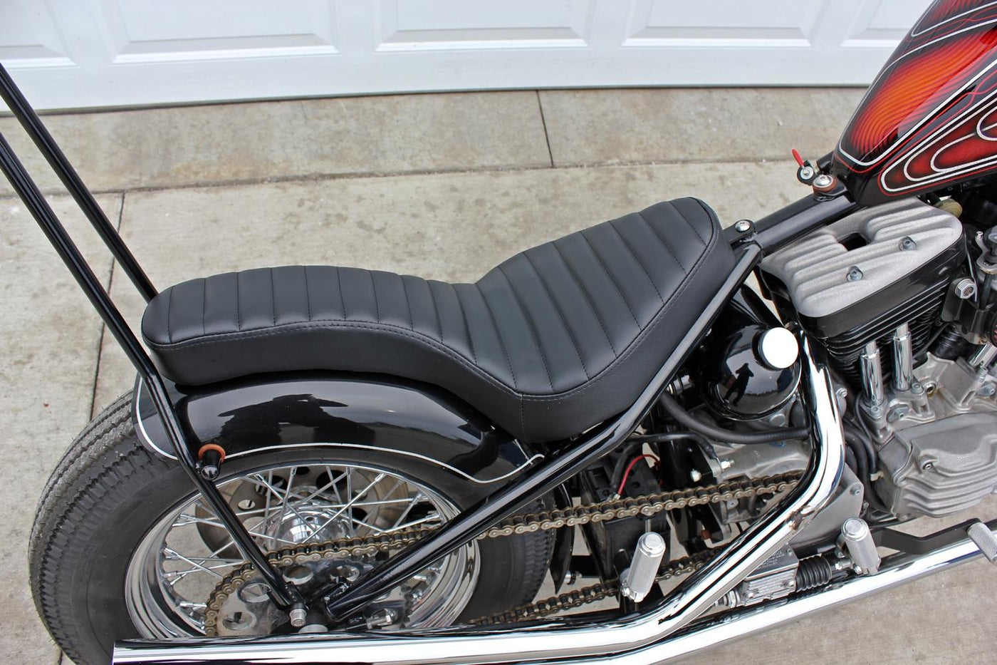 A motorcycle parked in front of a garage, featuring a TC Bros. Hardtail Rigid Cobra Seat Black Pleated.