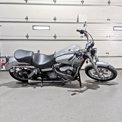 A silver TC Bros. Harley Davidson Dyna motorcycle, with an aftermarket exhaust system, parked in front of a garage.