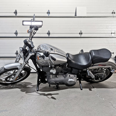 A silver TC Bros. Harley Davidson Dyna motorcycle parked in front of a garage with the TC Bros. Dyna Rear Crash Bar (fits 2006-2017 models) aftermarket exhaust system.