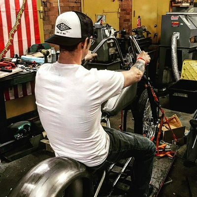 A man is working on a motorcycle in a workshop while wearing a TC Bros. Diamond Trucker Hat - White/Black with an Adjustable Snapback Closure.