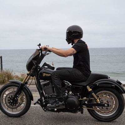 A man riding a black motorcycle near the ocean, equipped with a TC Bros. Harley Davidson Dyna Belt to Chain Conversion Kit (Chrome Sprocket) that fits 2006-17 Dyna Models.