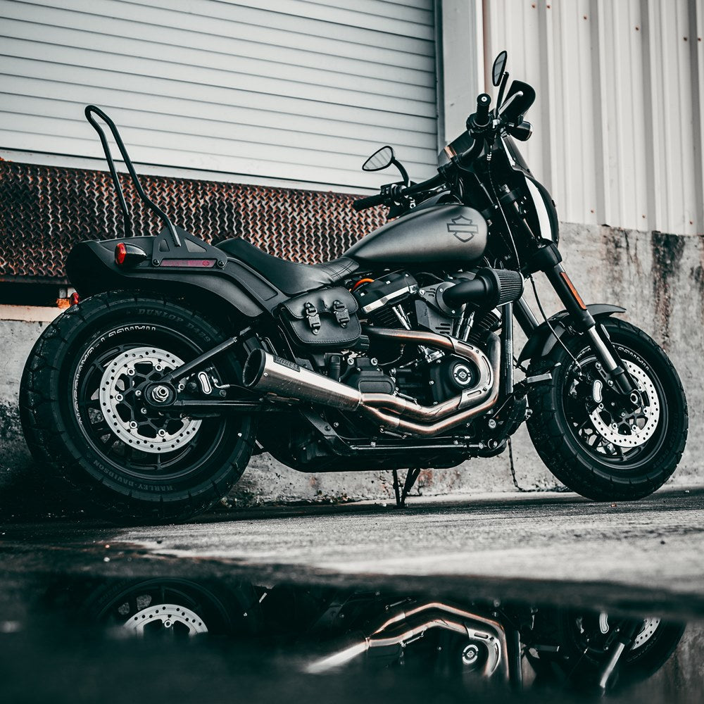 A black TC Bros. M8 Fat Bob Kickback Sissy Bar 2018-newer FXFB FXFBS motorcycle is parked in front of a building.