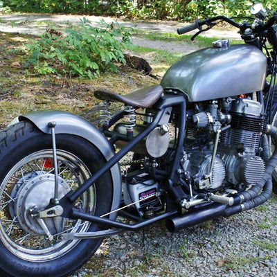 A TC Bros. vintage-style motorcycle parked in the woods with a 6-1/4" Wide Spun Steel Chopper & Bobber Fender by TC Bros Choppers.
