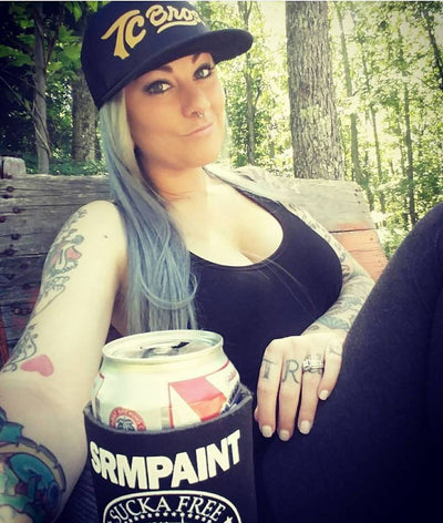 A woman with tattoos holding a can of beer and wearing a TC Bros. Script Trucker Hat - Black/Gold.