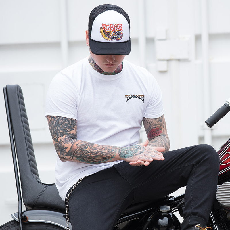 A man with tattoos sitting on a motorcycle wearing a TC Bros. Wing Trucker Hat - White/Black from TC Bros.