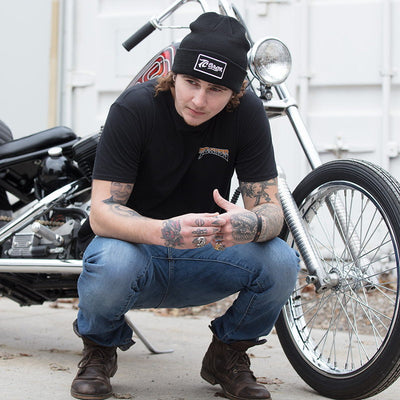 A man kneeling next to a TC Bros. motorcycle, wearing a TC Bros. Classic Beanie - Black and an Embroidered Patch.