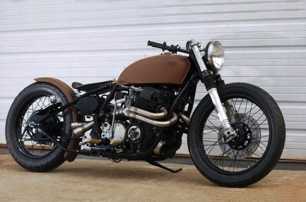 A brown TC Bros. Honda CB750 Weld On Hardtail Frame motorcycle parked in front of a garage, suitable for chopper or bobber enthusiasts.