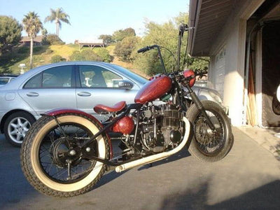 A red TC Bros. Honda CB750 Weld On Hardtail Frame motorcycle parked in front of a garage, creating a chopper or bobber aesthetic.