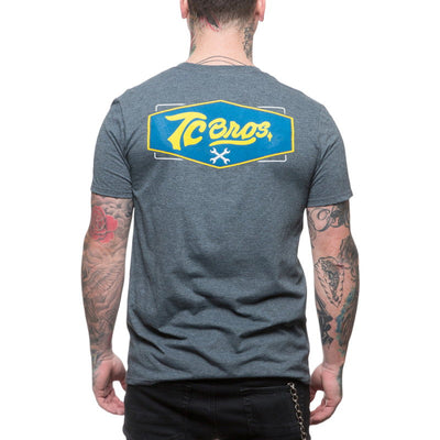 The back of a man wearing a TC Bros. Shield T-Shirt - Charcoal Heather with a yellow and blue logo.