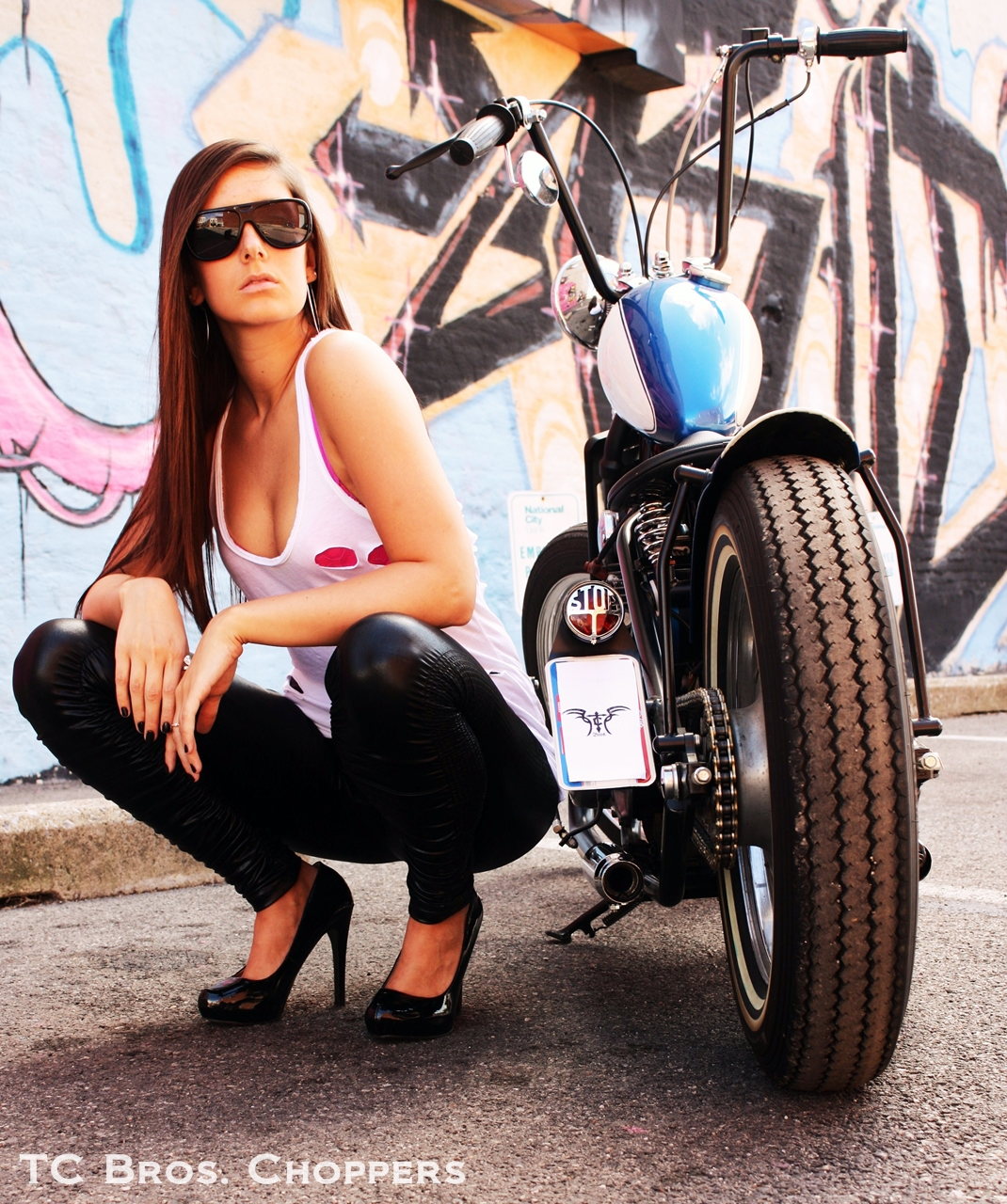 A woman posing in front of a motorcycle with TC Bros. 7/8" Ape Hanger Handlebars - 12" Black Powdercoated made of American steel tubing.