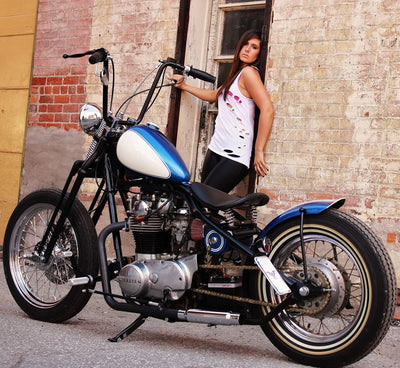 A woman standing next to a Shinko motorcycle in front of a brick wall with vintage style.