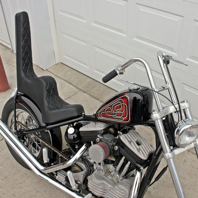 A black and silver TC Bros. motorcycle parked in front of a garage, featuring TC Bros. King & Queen Hardtail Seat with black diamond pleated stitching.