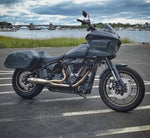 The SP Concepts Lane Splitter Exhaust for M8 Softail with Factory Bags in a stainless finish is displayed on a workbench, with visible welds and a welding helmet featuring an American flag design in the background.