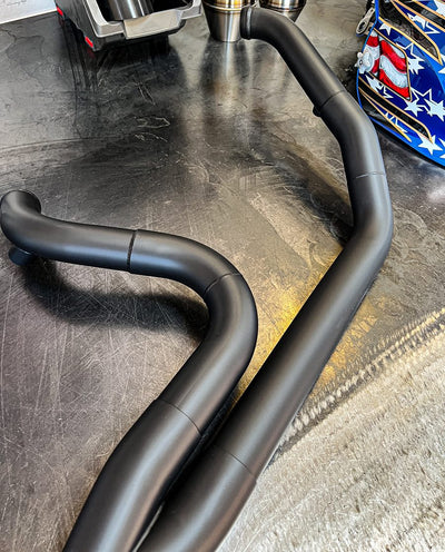 A pair of SP Concepts Big Bore Exhaust M8 Touring FLT 2017-Present (black) mufflers on a table in a garage, featuring high flow performance rolled cone muffler.