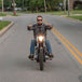 A man riding a motorcycle down a street with the Triangle Chopper Headlight - Aris Style -Chrome from Moto Iron® illuminating his way.